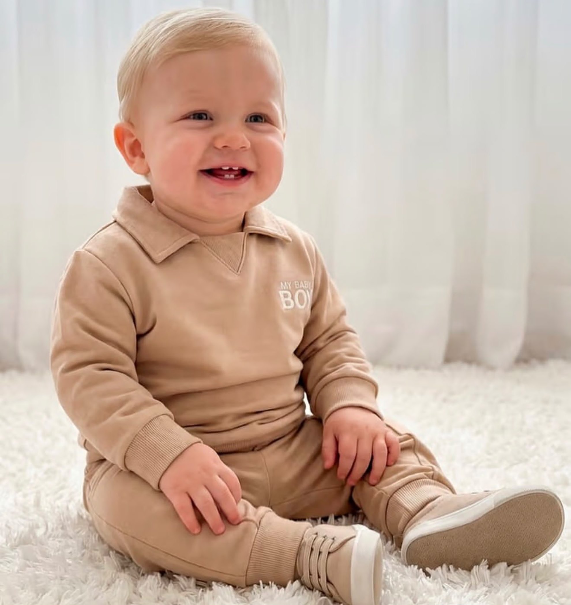 Bebê sorrindo sentado com conjunto de moletom bege e tênis